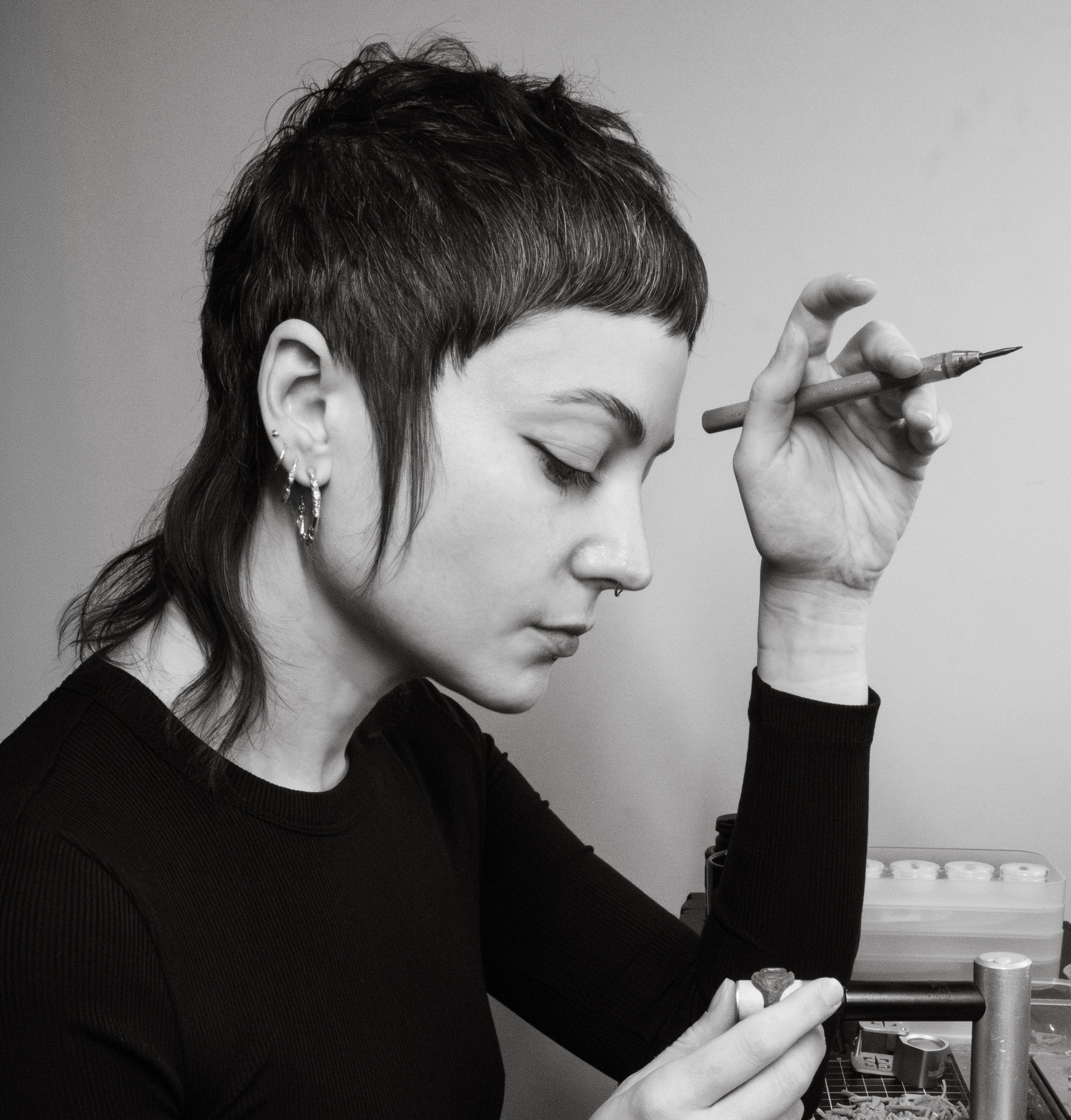 Profile photo of jeweller Alicia Hannah Naomi, at her workbench. She is staring down at a wax model she is carving, holding a wax carving tool in her opposite hand.