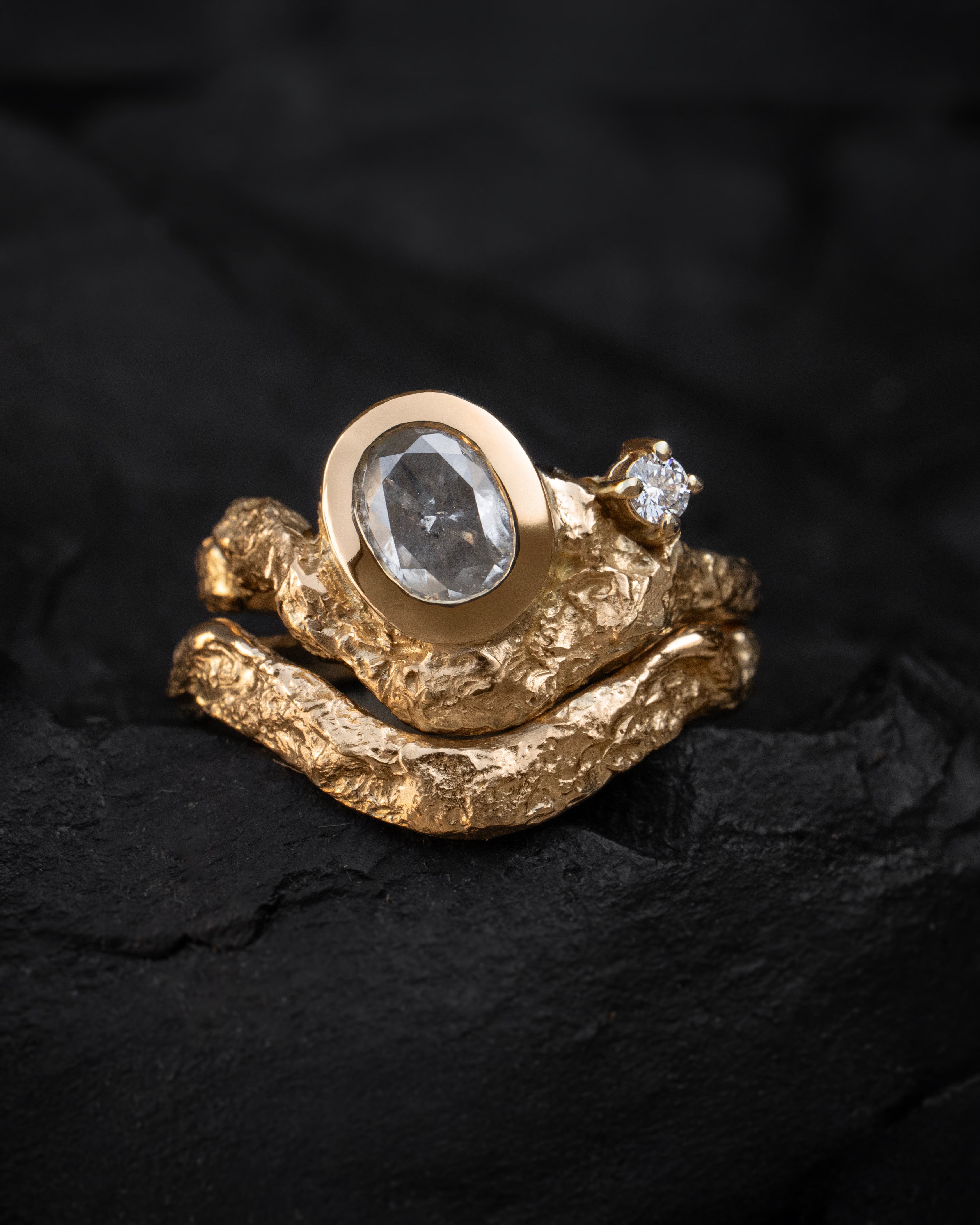Sculptural gold engagement ring with a bezel set icy salt and pepper diamond central stone and smaller white diamond accent on a dark background.