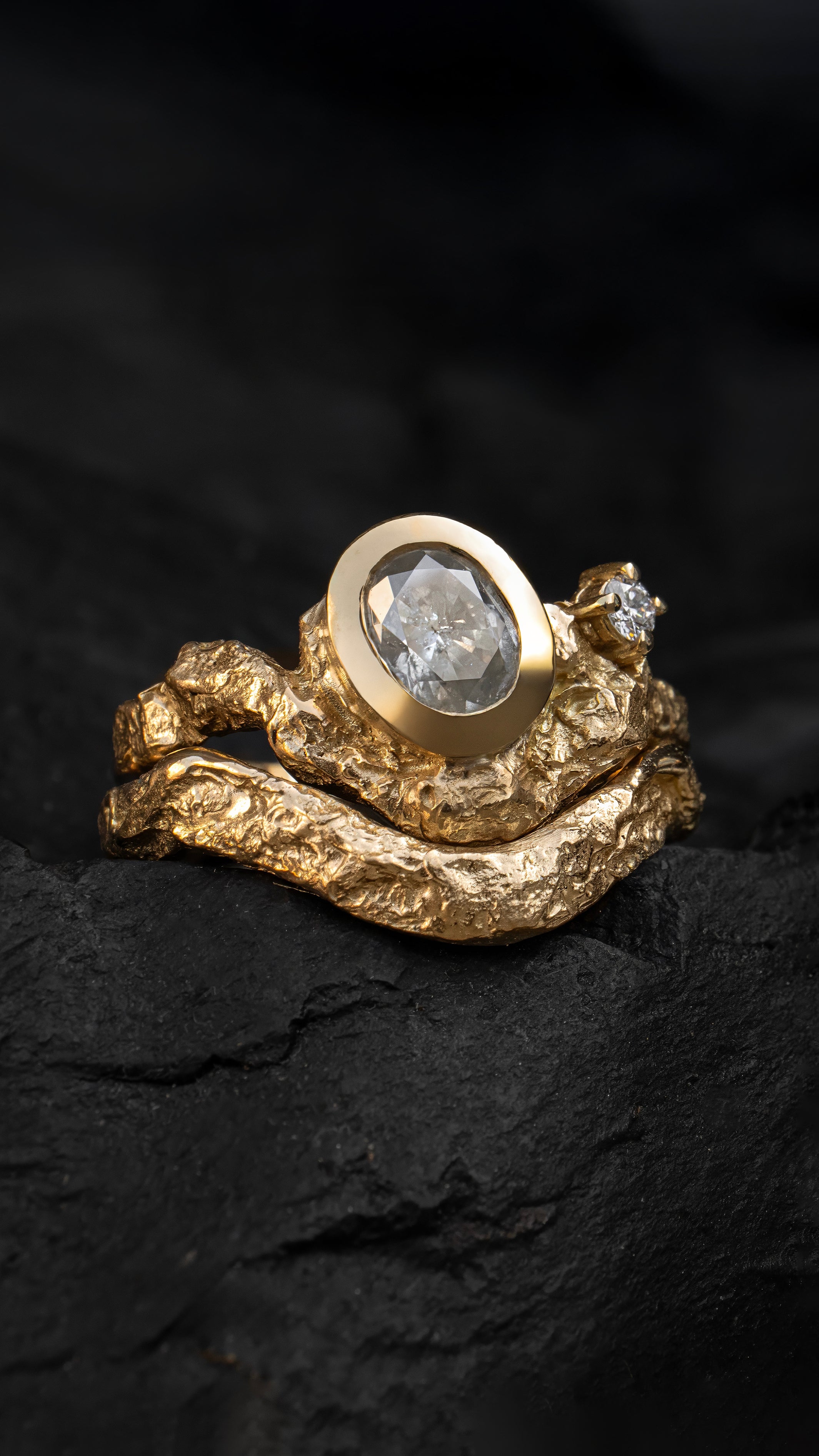 18ct Yellow Gold Alternative Engagement Ring with rugged texture and smoky white diamonds stacked with a matching wedding band on a black fabric background