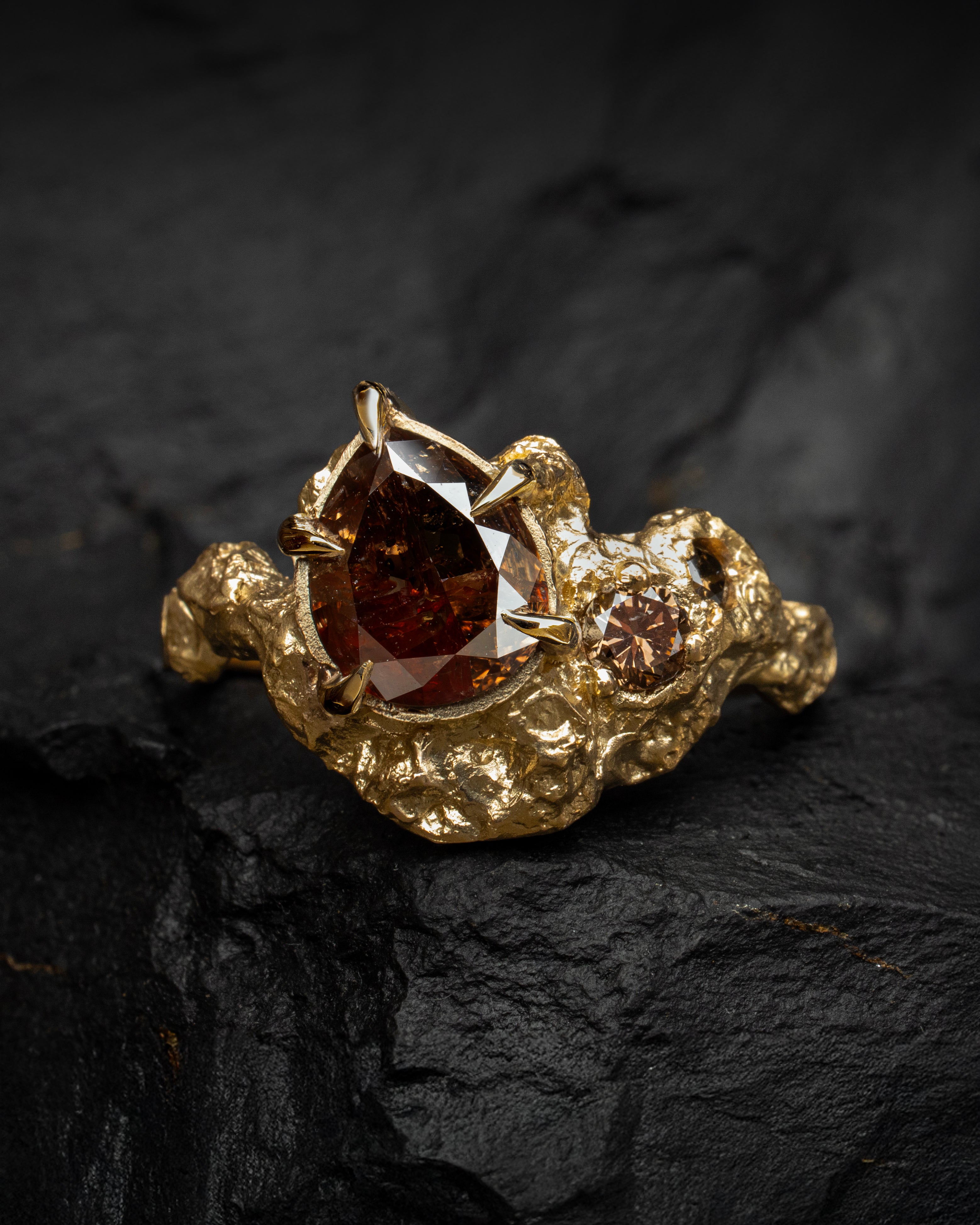 Sculptural gold engagement ring with a large brown diamond central gemstone and champagne diamond accents with lots of texture on a dark stone surface.