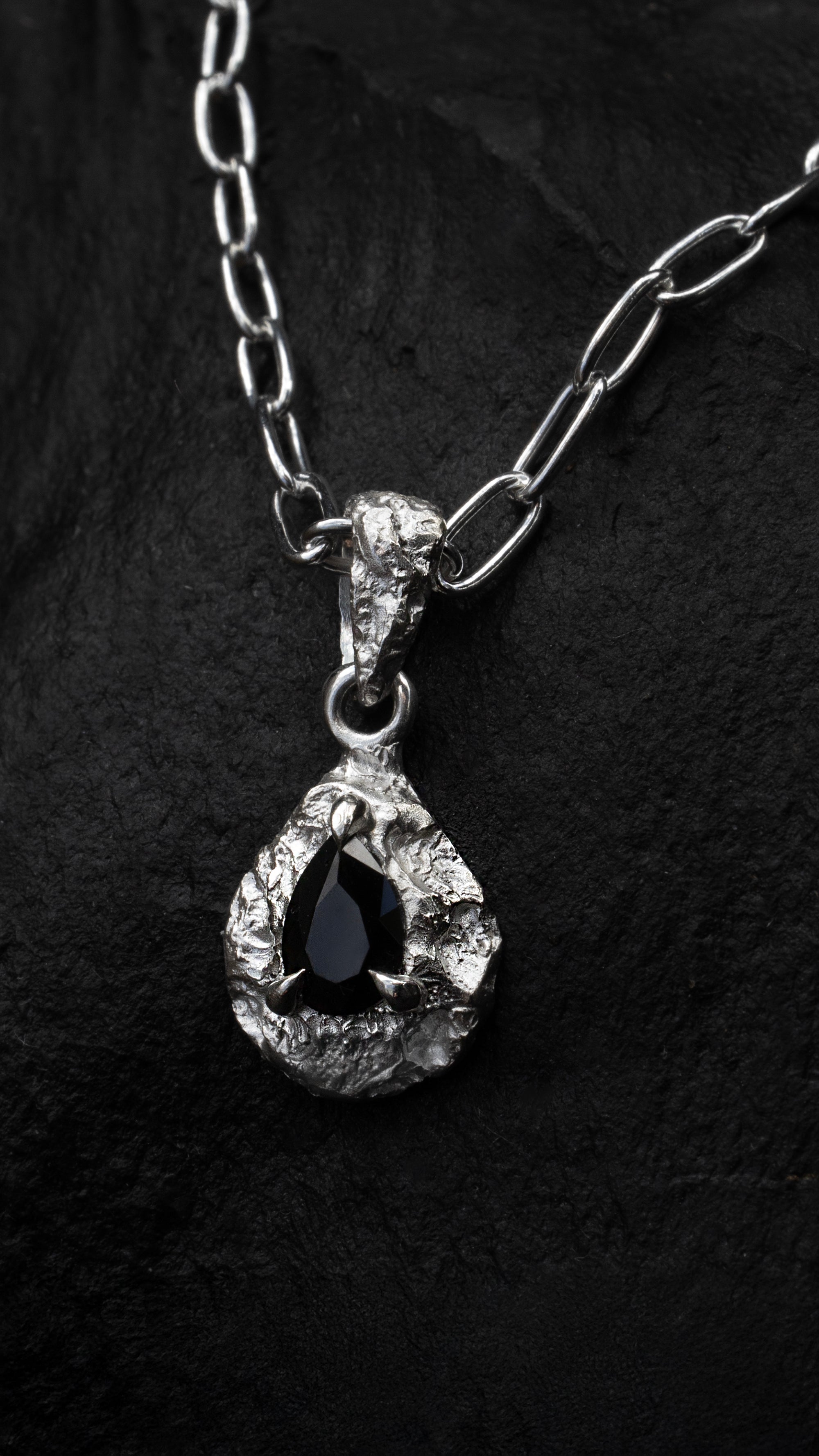 Ruggedly textured teardrop shaped silver pendant with a pear-cut black gemstone set in three claws at the centre. Pictured against a black rock.