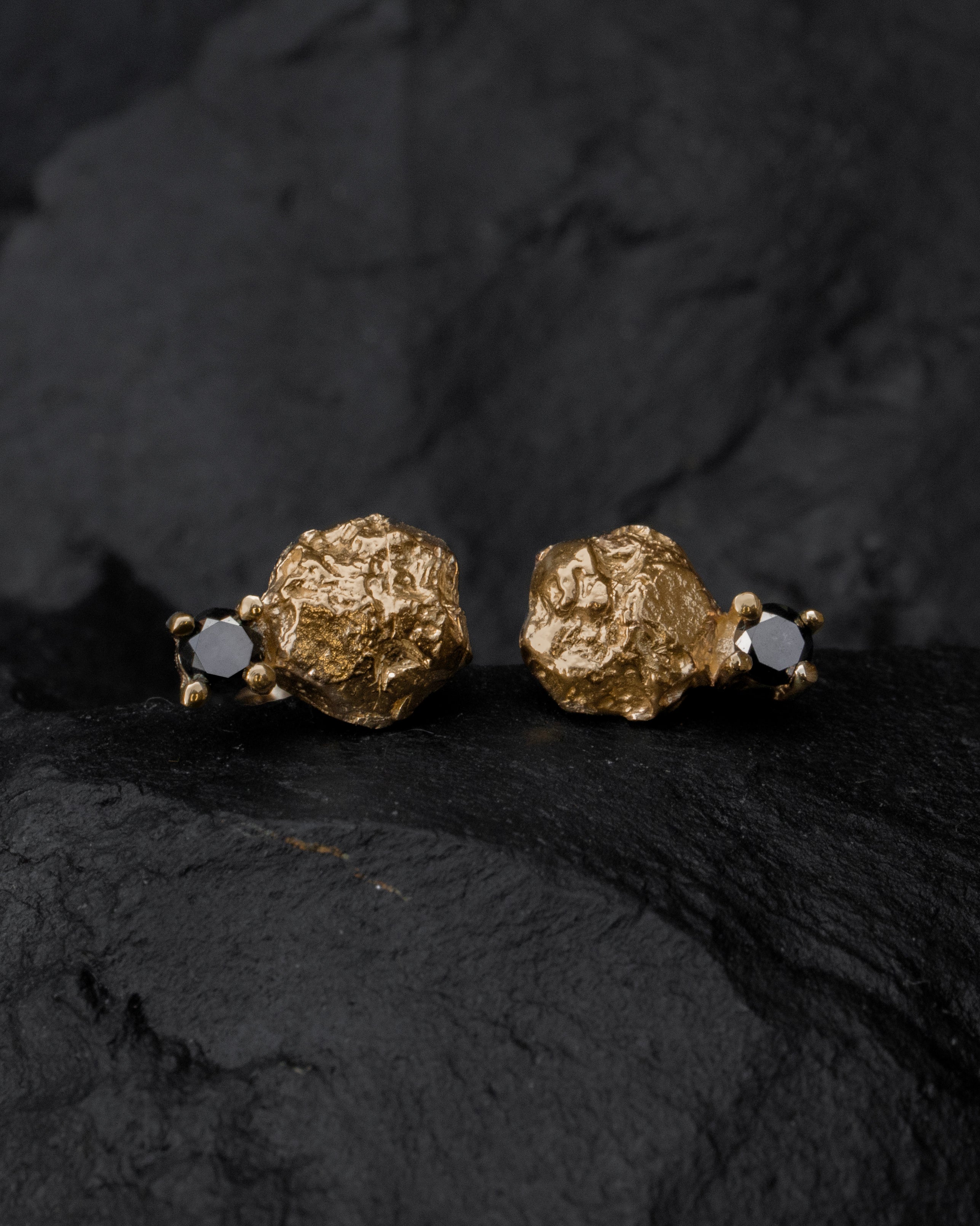 Small pebble-like yellow gold stud earrings with black diamond accent stones, pictured on a black background.