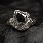 A gothic-style ring featuring a large, faceted black pear cut diamond in a textured, 18ct white goldsetting, resting on a dark, rough stone surface.