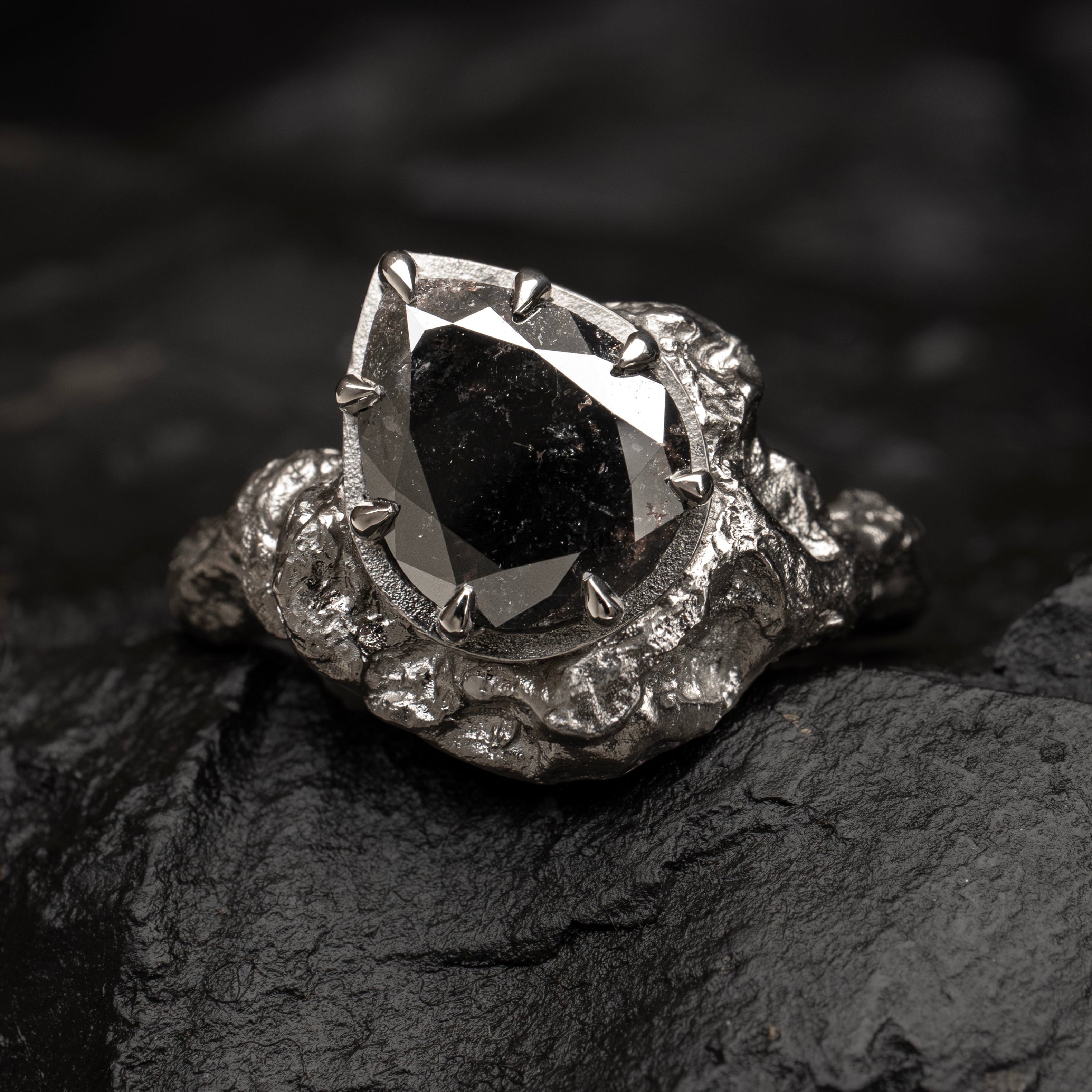 A gothic-style ring featuring a large, faceted black pear cut diamond in a textured, 18ct white goldsetting, resting on a dark, rough stone surface.