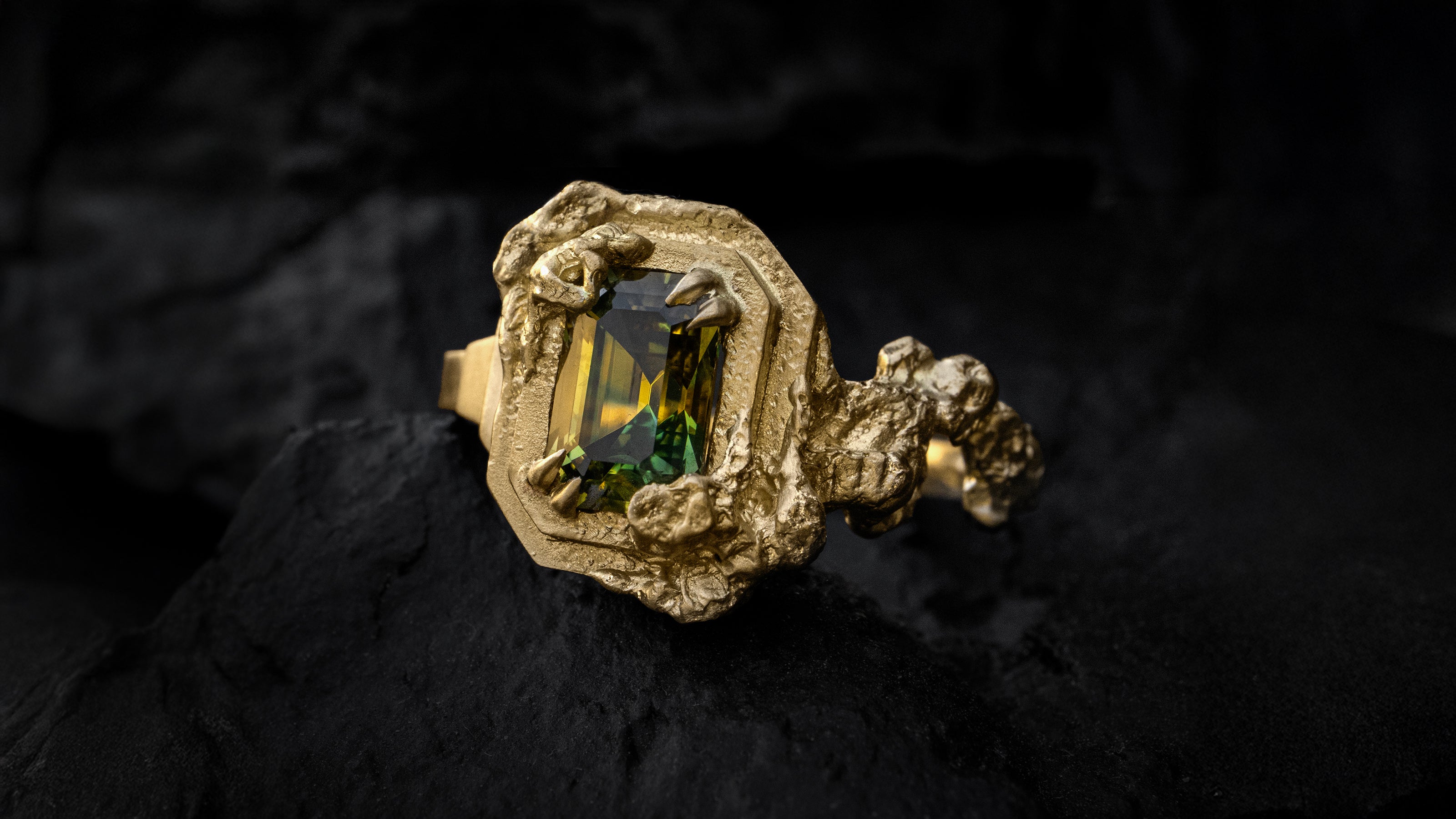 A beautiful Art Deco inspired engagement ring made of 18ct Yellow Gold with encroaching cloud-like texture surrounding the central Australian parti-sapphire emerald cut gemstone. Pictured on a black background.